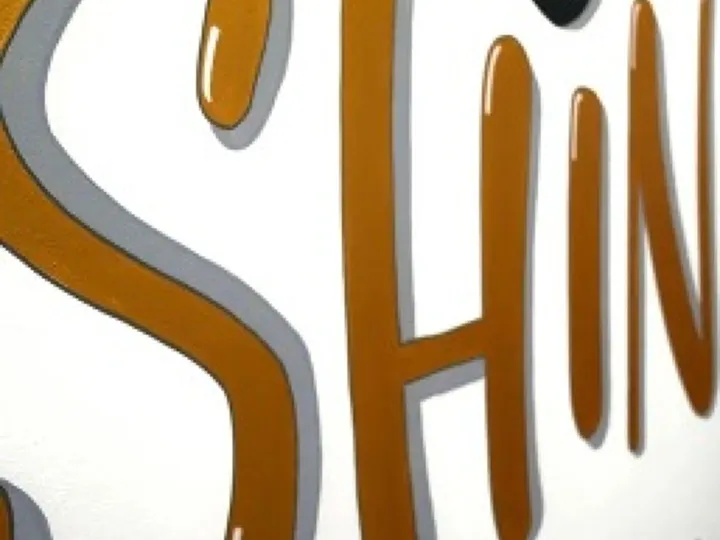 The word Shine hand lettered in gold on white board