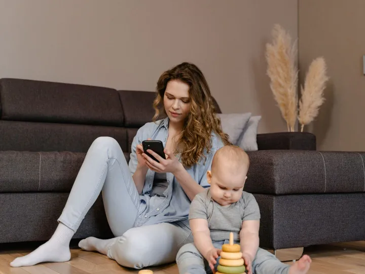 Parent sitting on the floor searching on her phone while her baby plays with stacking rings