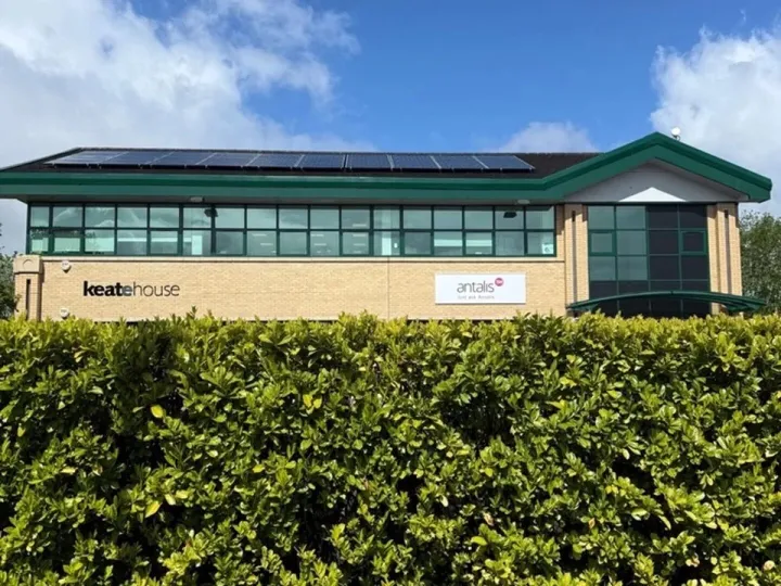 Brick office building with green window frames and rooftop solar panels behind a tall hedge