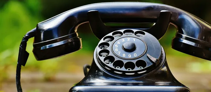 Close-up of a classic black rotary phone outdoors with a blurred green background.
