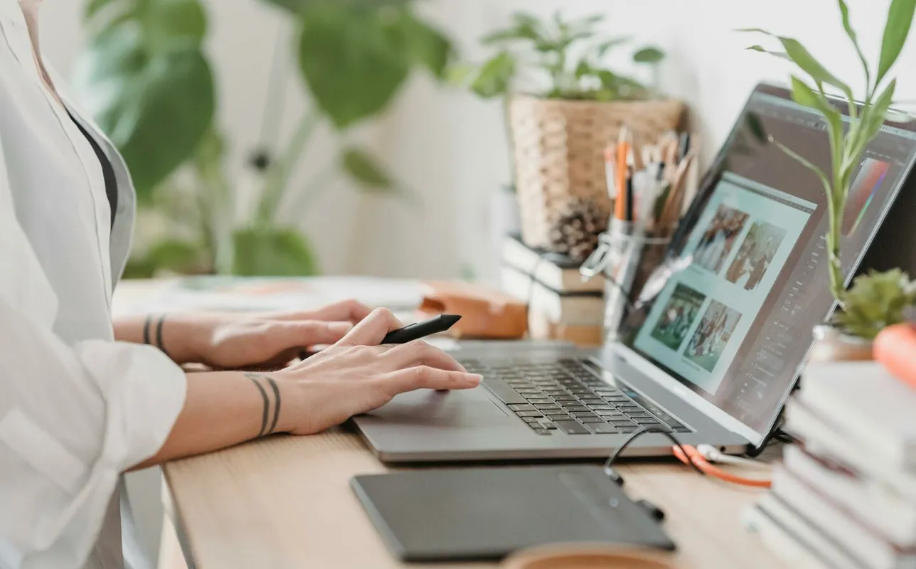 Freelance designer working from home using a laptop and digital tablet, surrounded by plants.