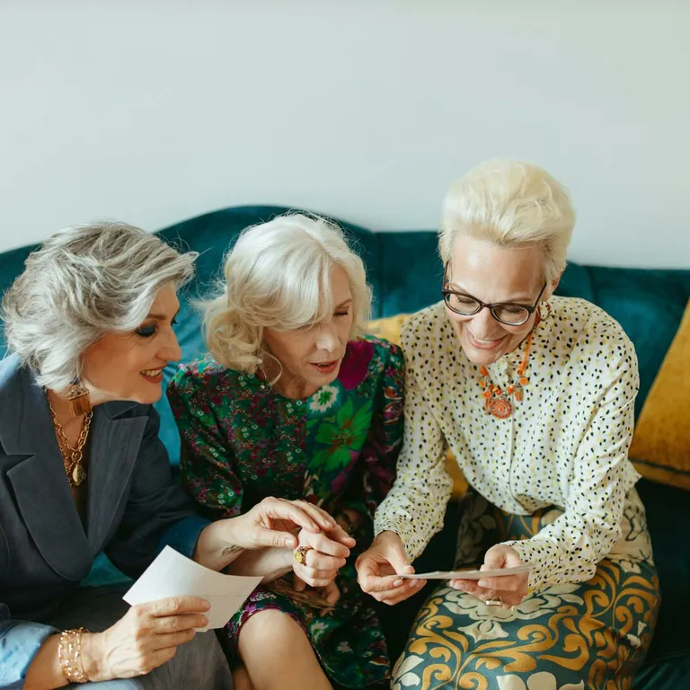 Three senior women joyfully reminiscing over old photos on a cozy couch.