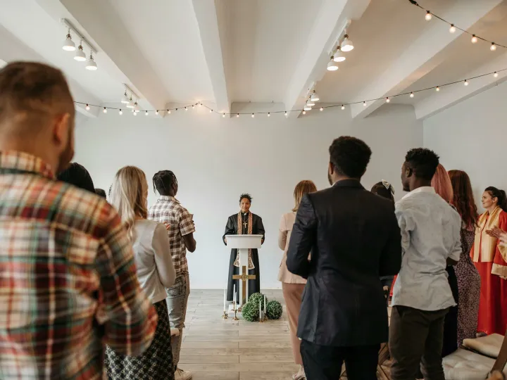 A diverse congregation stands together in prayer led by a woman pastor indoors.