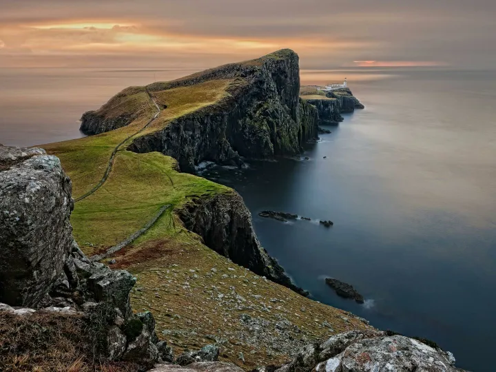 A stunning sunset view of Neist Point Lighthouse with cliffs and serene sea in Scotland.