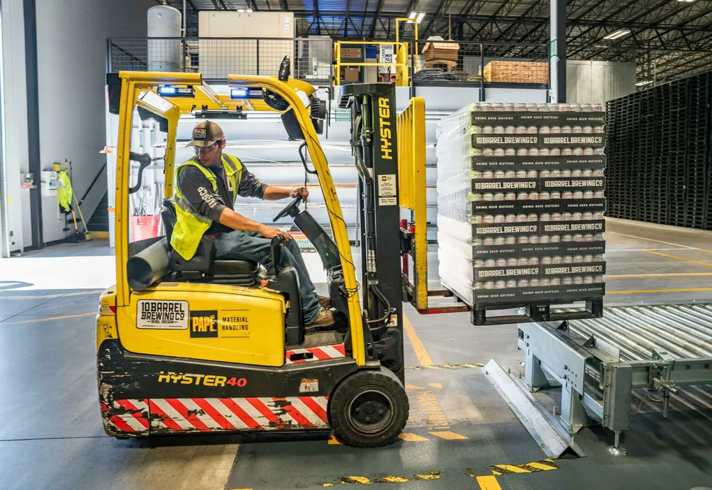 A warehouse worker maneuvers a forklift to transport crates for brewing company storage.