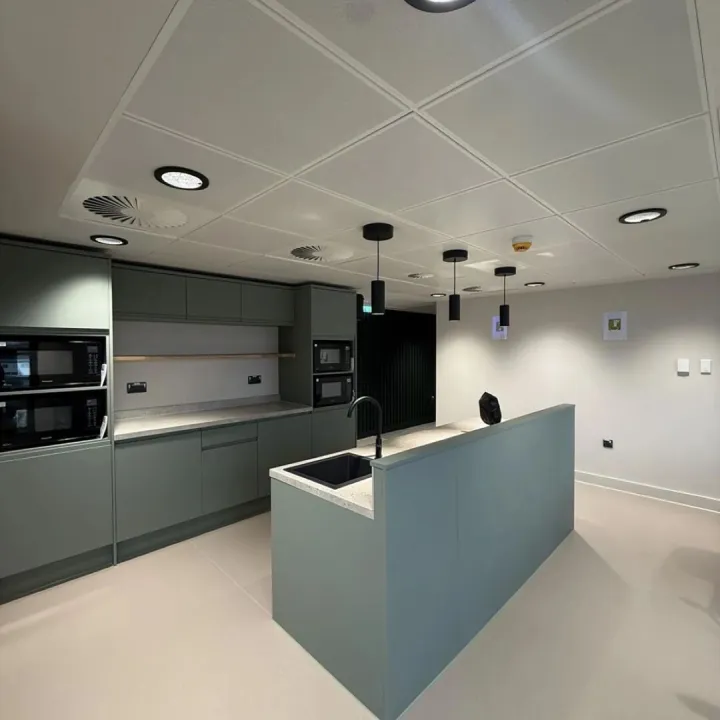 Office kitchen with sage green units, built-in microwaves, an island sink and black pendant lights
