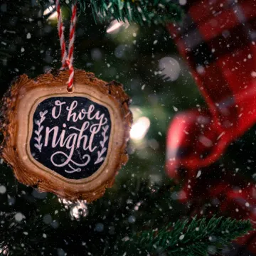 Wooden bauble with O Holy Night inscription hanging on green Christmas tree decorated with red ribbon on snowy day