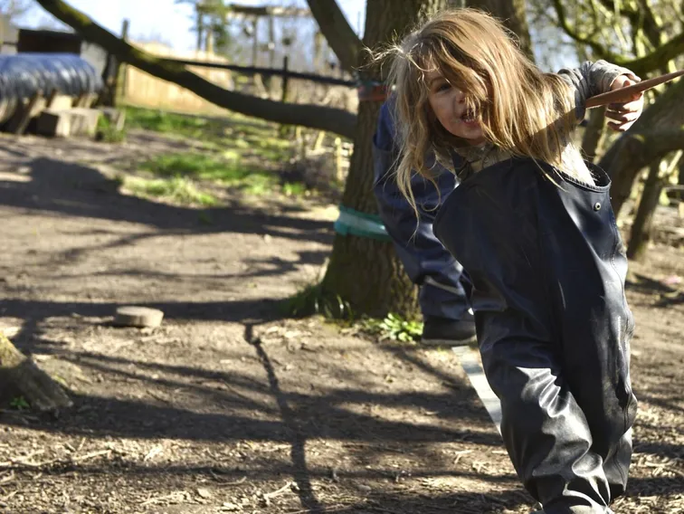 A child in waterproofs balancing along a slackline between the trees, holding the guide rope