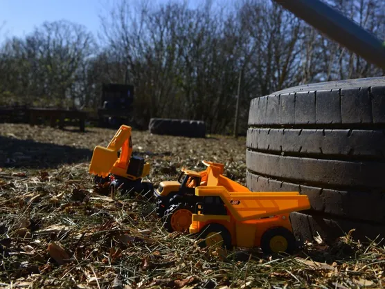 Toy diggers and a dump truck in the woodchip beside a big tyre, with trees behind