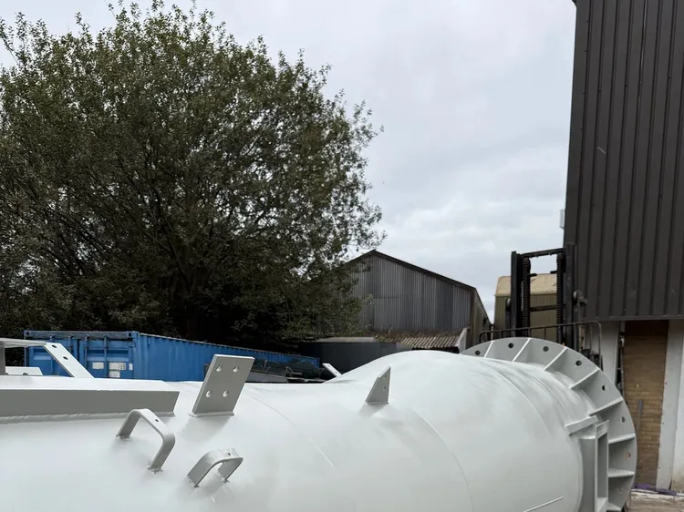 Primed grey steel chimney section with lifting lugs and a stiffened flange end, outside a workshop