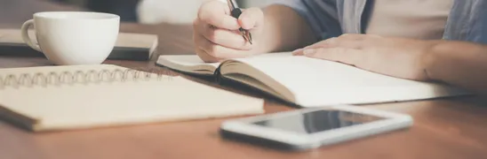 Woman Writing on a Notebook Beside Teacup and Tablet Computer