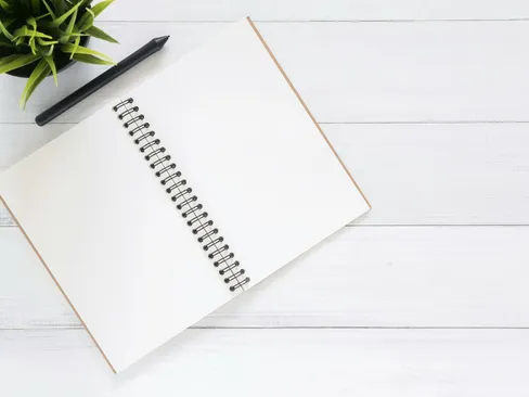 Open spiral notebook on white desk with black pen and green plant. Ideal for planning.