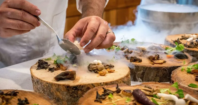 A chef artfully plating a gourmet dish with mushrooms and greens on wood slices.