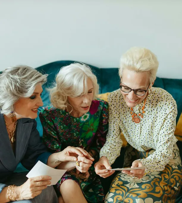 Three senior women joyfully reminiscing over old photos on a cozy couch.