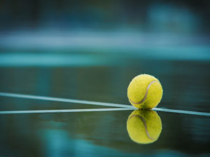 A tennis ball sits on a wet court with its reflection visible, capturing a serene sports moment.