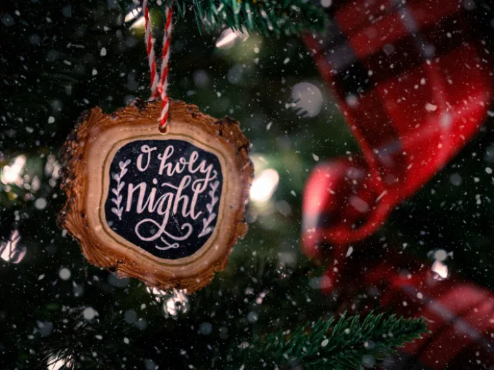 Wooden bauble with O Holy Night inscription hanging on green Christmas tree decorated with red ribbon on snowy day