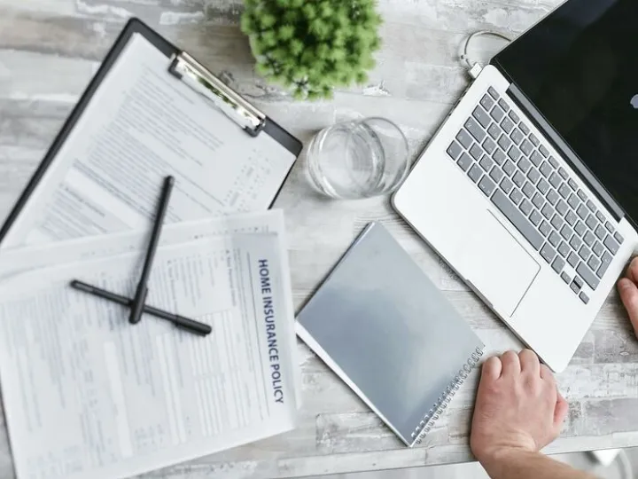 Person Writing on White Paper Beside Clear Glass Mug and Silver Macbook